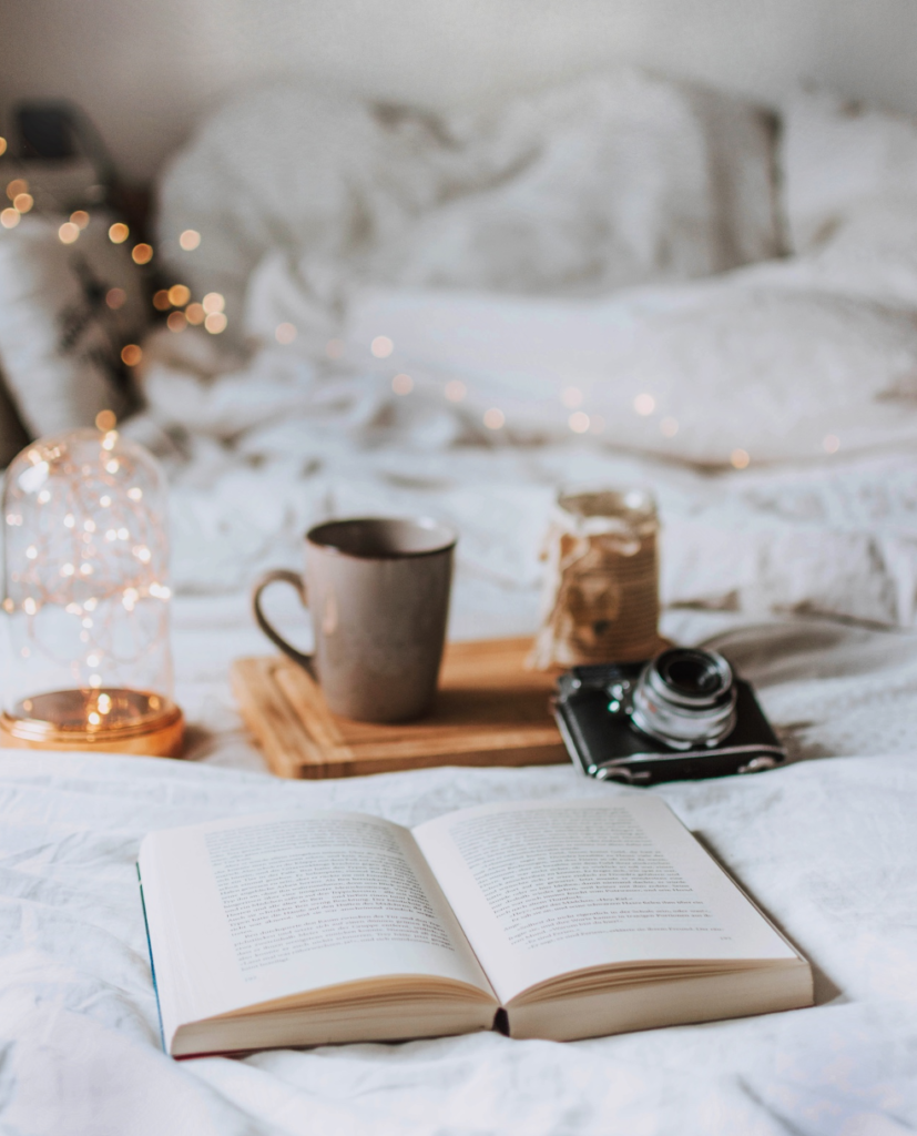 postel se světýlky v pozadí, vpředu hrnek s kávou a rozložená kniha